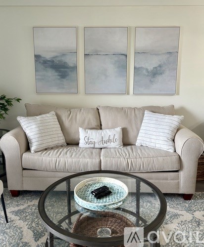 A living room with a beige couch, a glass coffee table, and three framed artworks on the wall.