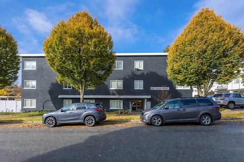 Two cars are parked in front of a grey building with trees.