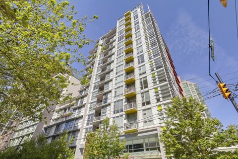 A tall building with many windows and balconies is surrounded by trees and has a clear blue sky in the background.