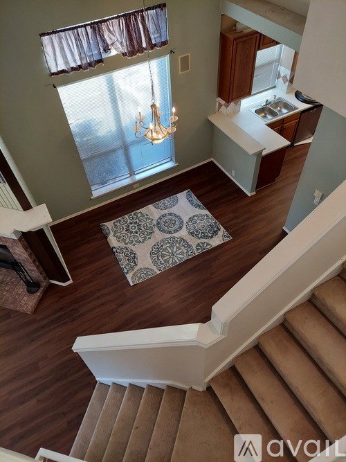 A chandelier hangs over a patterned rug in a well-lit room with wooden floors and stairs.