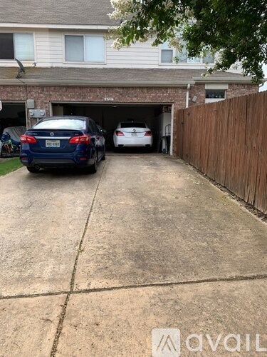 A dark blue sedan is parked in a driveway with a white car in the garage.