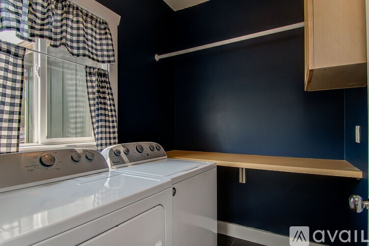 A washing machine sits in a laundry room with a window and checkered curtains.
