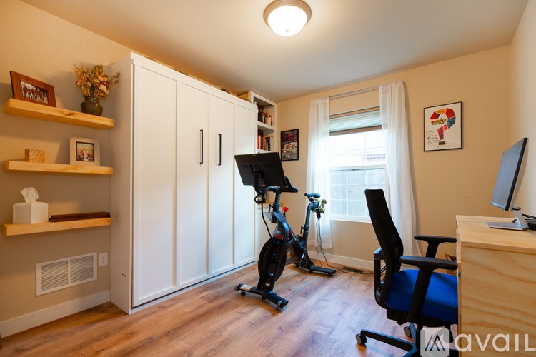 A room with a wooden floor, a desk with a computer, a chair, a bicycle, and a window with white curtains.