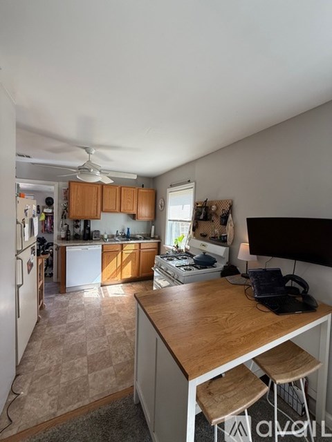 A kitchen with a refrigerator, a table with a laptop and a fan on the ceiling.