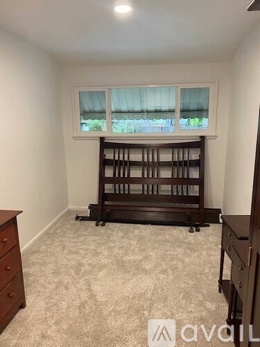 A room with a carpeted floor, a window with blinds, and a wooden bookshelf.