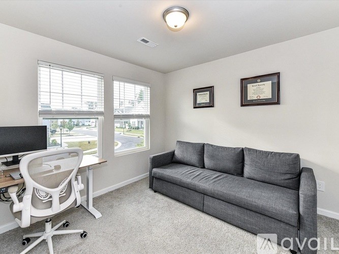 A living room with a grey couch, a desk with a computer, and two framed certificates on the wall.