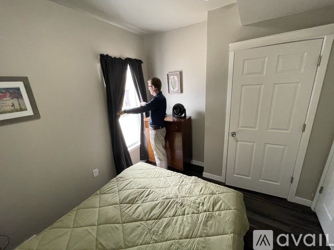 A man is opening curtains in a room with a bed and a dresser.