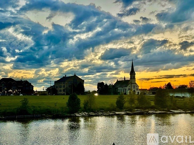 A church with a tall steeple stands in front of a house by a body of water.