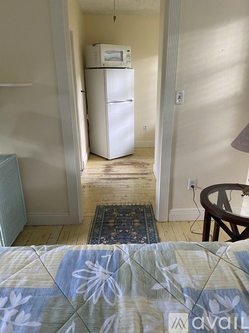 A bedroom with a bed, a chair, and a refrigerator in the hallway.
