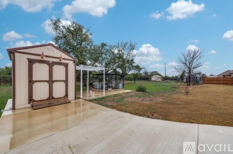 A small wooden structure with a brown roof is situated on a concrete slab with a grassy area and trees in the background.