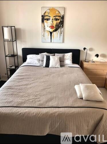 A bed with a grey comforter and a white pillow is in the foreground with a painting of a woman's face on the wall behind it.