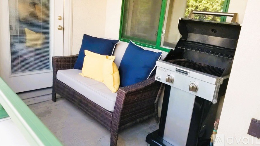 A black grill is on a patio next to a couch with blue and yellow pillows.