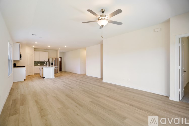 A spacious room with a ceiling fan and light, hardwood floors, and a kitchen area with white cabinets and appliances.