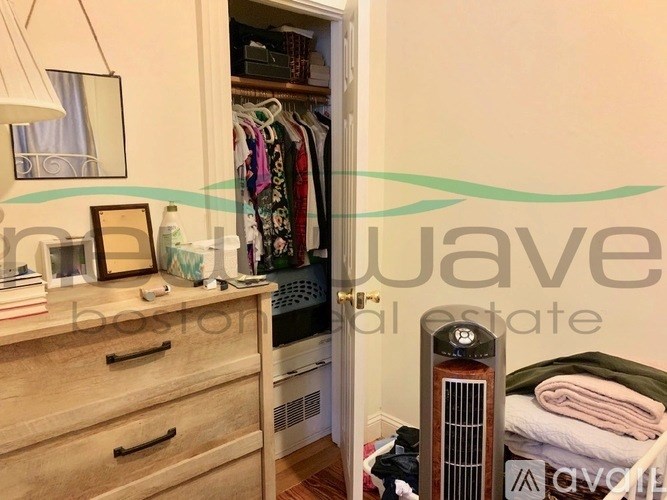 A room with a wooden dresser, a mirror, a lamp, a closet with clothes, a radiator, and a bed with towels.