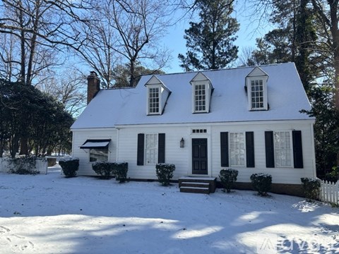 A white house with black shutters and a black door is surrounded by snow.