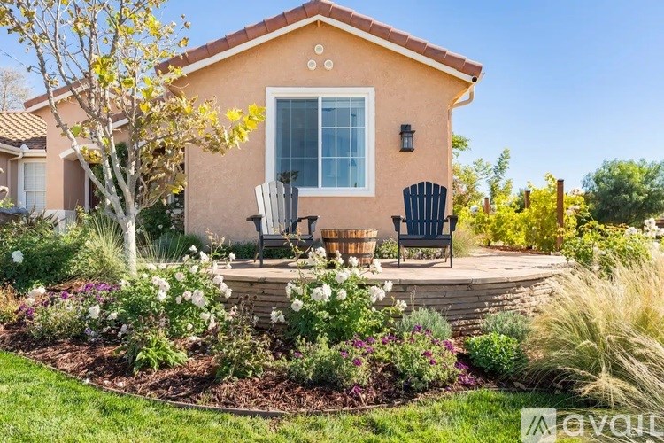 A house with a brown roof and a white window is surrounded by a garden with a black chair and a table.