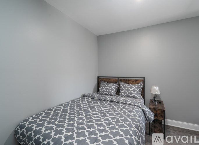A bed with a grey and white patterned comforter and two matching pillows sits in a room with a wooden headboard and a small table with a lamp.
