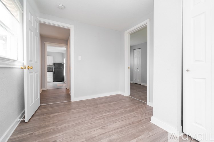A room with white walls and wooden flooring, with a door on the right and a hallway leading to another room.