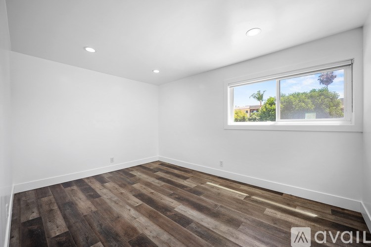 A room with wooden flooring and a window showing a view of trees and a building outside.
