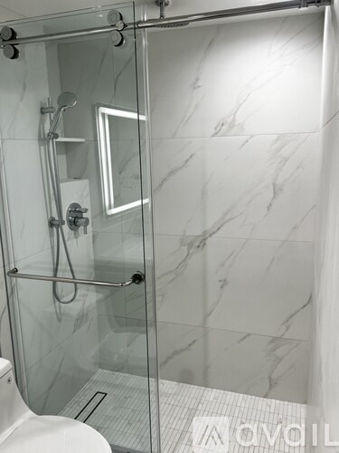 A white toilet is in a marble bathroom with a walk-in shower.