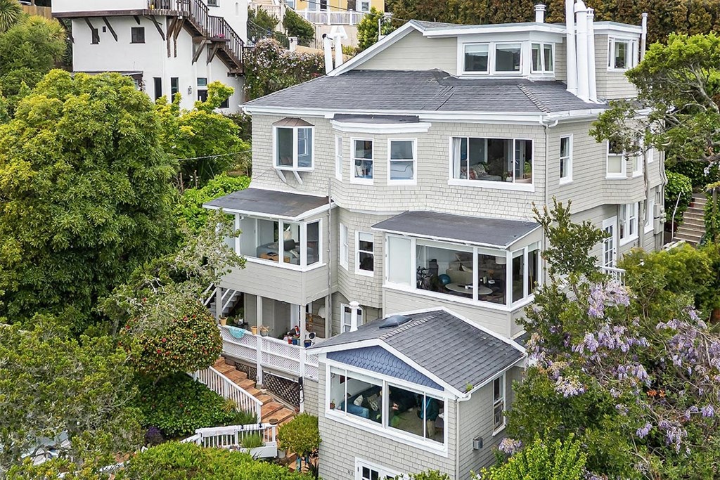 A large, multi-level house with a grey roof and white walls is surrounded by greenery.
