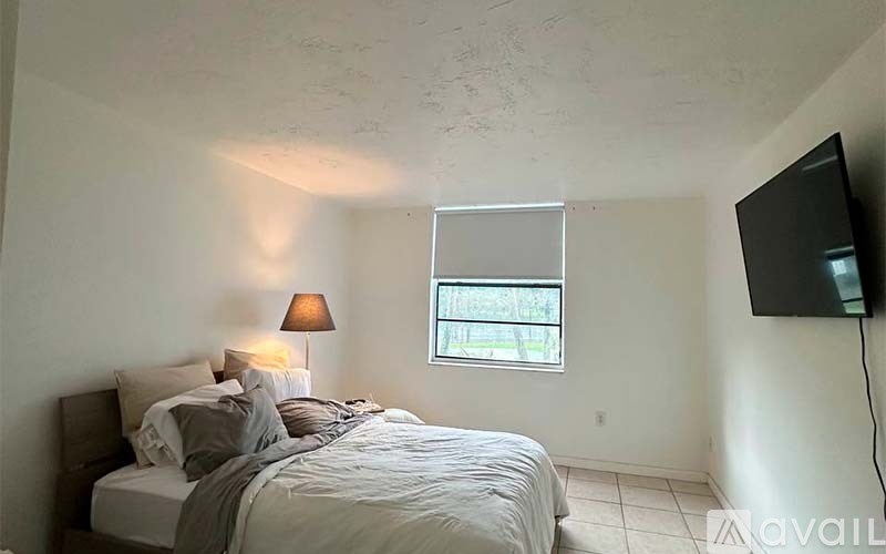 A bedroom with a bed, a lamp, a window, and a flat screen TV mounted on the wall.