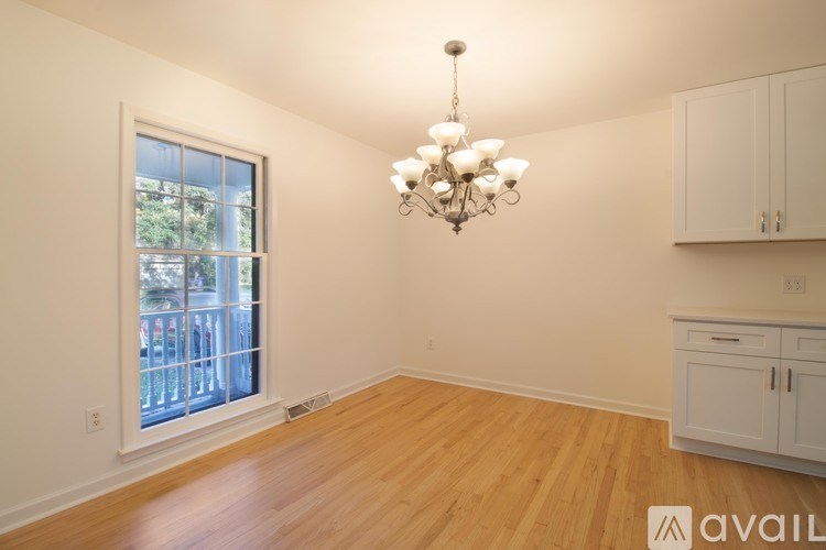 A room with wooden floors, a chandelier, and white walls with a window and cabinets.