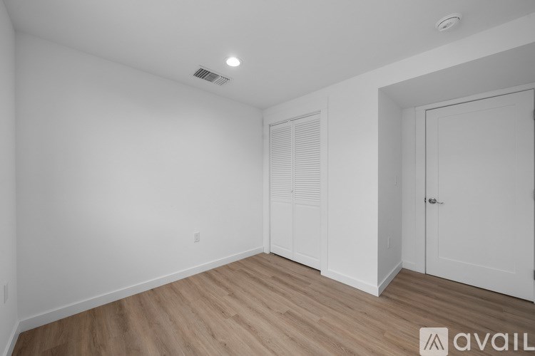 A room with wooden flooring and white walls, with a door on the right and a window with blinds on the left.