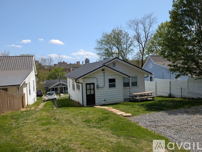 A small white house with a black door and windows is surrounded by a gravel driveway and a grassy yard.