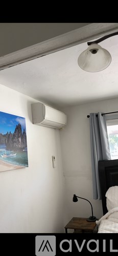 A room with a bed, a nightstand with a lamp, a painting on the wall, and an air conditioner.