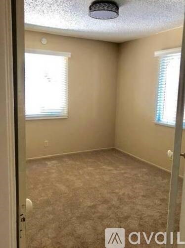 A room with beige walls and carpeting, a window with blinds, and a ceiling fan.