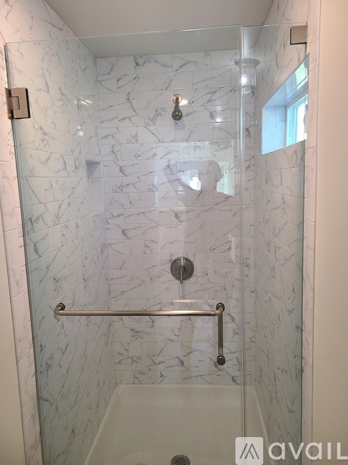 A white marble tiled shower with a glass door and a silver grab bar.