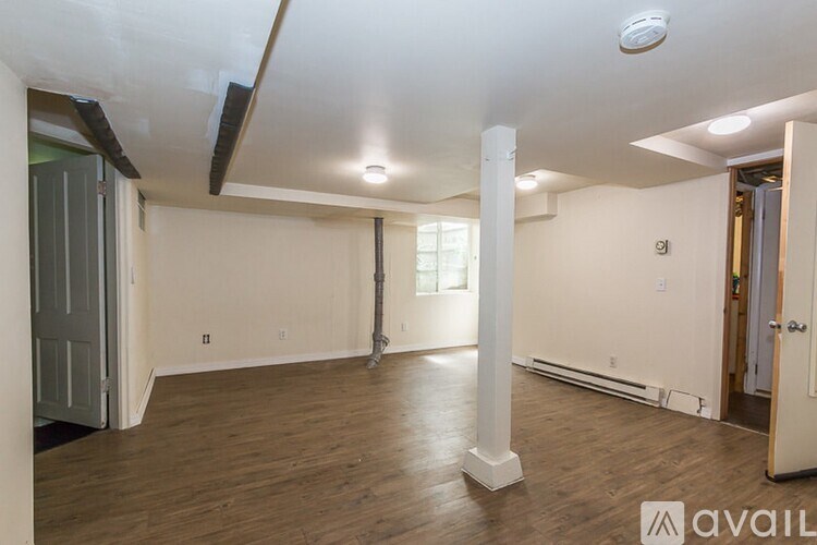 A spacious room with wooden floors and white walls, featuring a large window and a door on the left side.