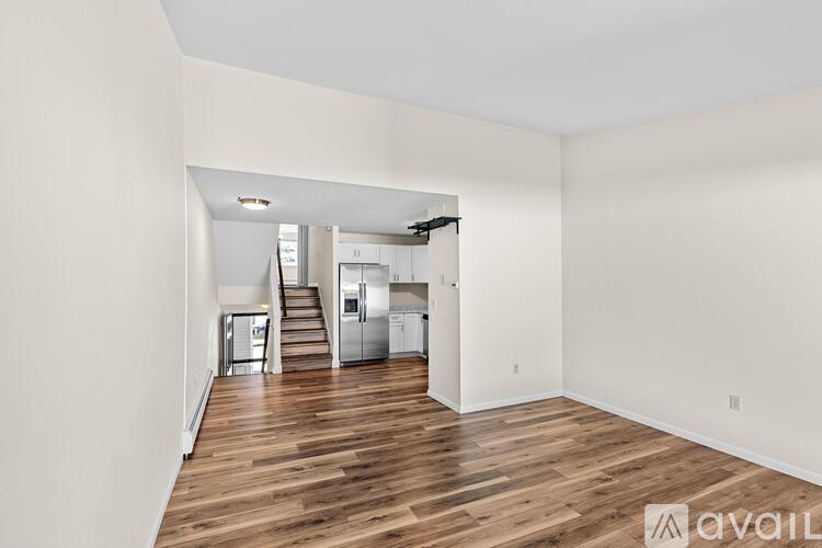 A spacious room with wooden floors and white walls, leading to a kitchen area.