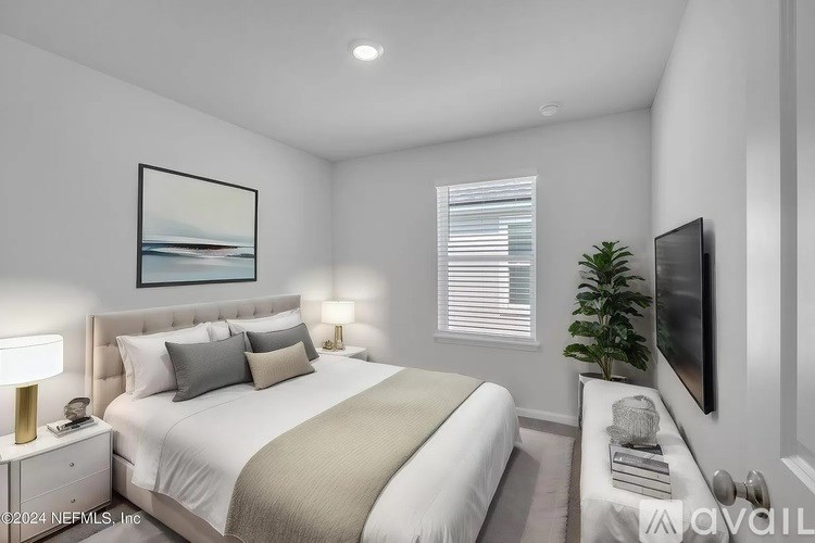 A bedroom with a large bed, a nightstand, a lamp, a plant, and a painting on the wall.