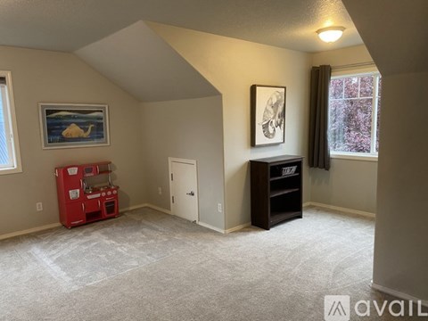 A room with a carpeted floor, a window with a view of trees, a white door, a red toy kitchen, a black shelf, and a painting on the wall.