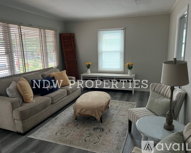 A living room with a couch, ottoman, and a table with a lamp on it.