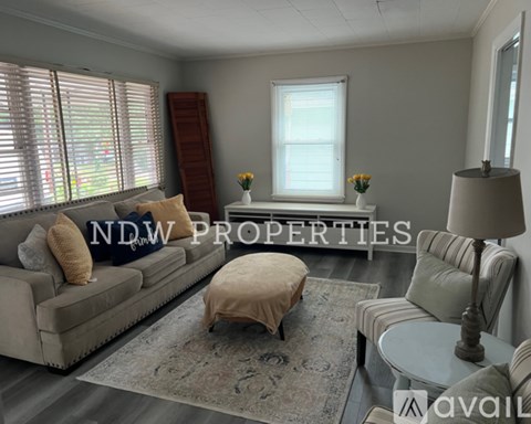 A living room with a couch, ottoman, and a table with a lamp on it.