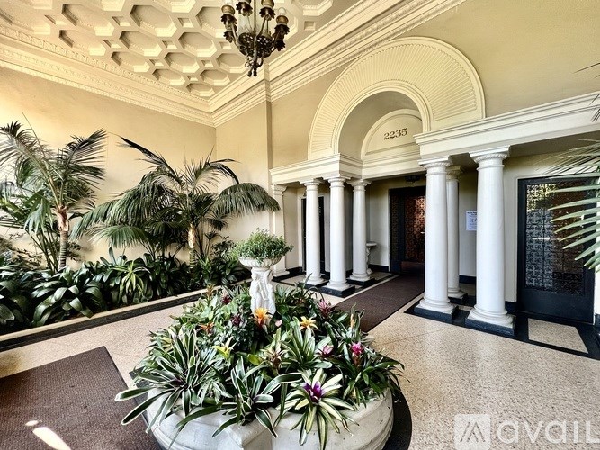 A large white vase with green plants in it sits on a brown rug in front of a row of white columns.