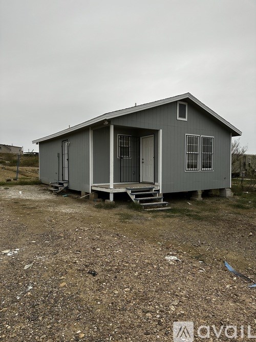 A small grey house with a porch and a door is situated in a gravel area.