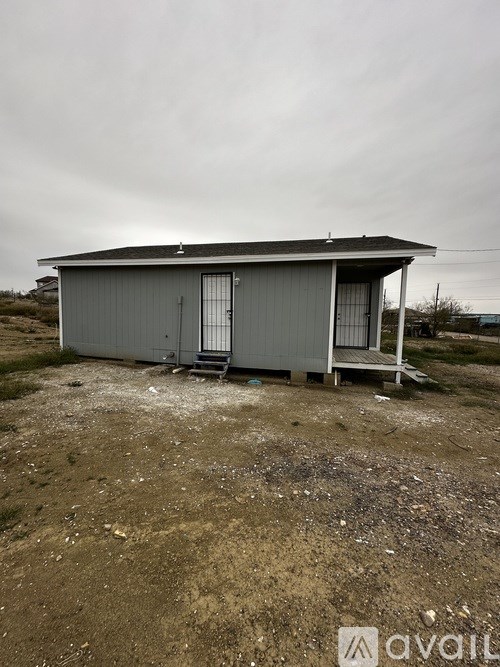 A grey metal shed with a black roof and a white door is situated on a dirt ground.