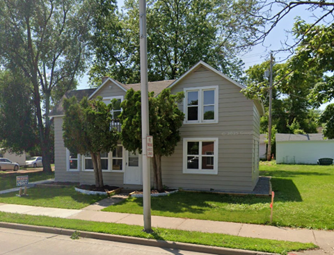 A house with a grey siding and a white door is surrounded by greenery.