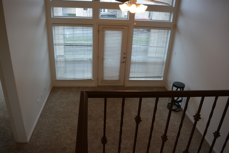 A room with a carpeted floor, a window with blinds, a chandelier, and a staircase with a railing.