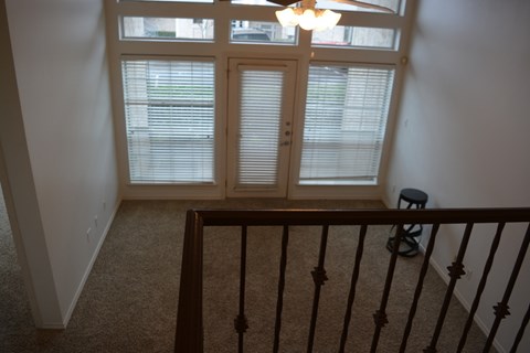 A room with a carpeted floor, a window with blinds, a chandelier, and a staircase with a railing.
