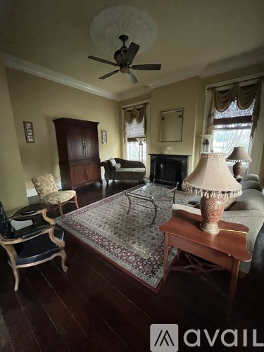 A living room with a rug, a couch, a chair, a table with a lamp and a fan on the ceiling.