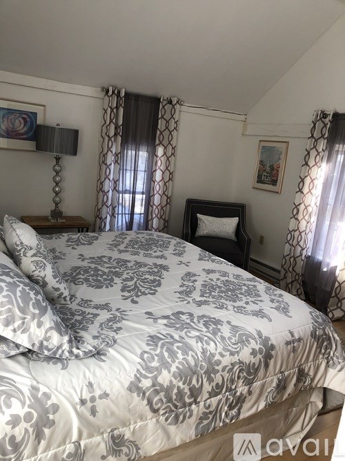 A bed with a grey and white patterned comforter is in the foreground of a room with a chair and a table with a lamp.