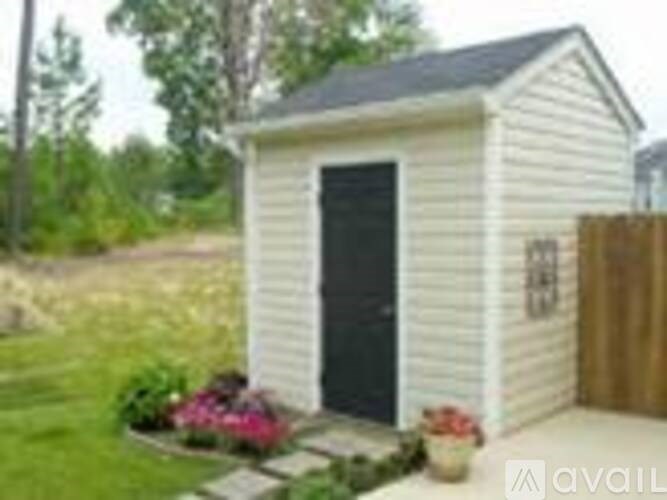 A small white shed with a black door is surrounded by a wooden fence and some flowers.