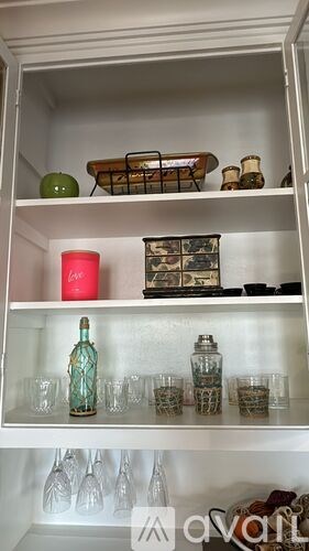 A white cabinet with a green apple, a pink candle, and a collection of glasses and bottles on the shelves.
