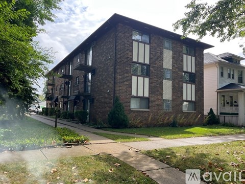 A brick building with a balcony on the second floor is surrounded by greenery and other buildings.