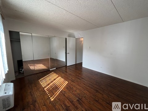 A room with wooden floors and white walls, with a mirrored wall and a window letting in sunlight.